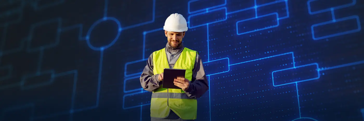 A person wearing a hard hat and high‑visibility vest stands in front of a digital flowchart display while using a tablet.